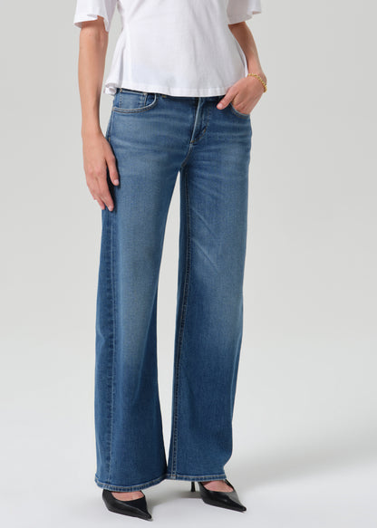 A model wearing a pair of wide-leg blue denim jeans, styled with a white top and black pointed-toe heels, photographed against a grey background.