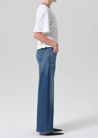 A side-profile view of a model wearing blue wide-leg jeans, a white short-sleeve t-shirt, and black pointed shoes against a grey background.