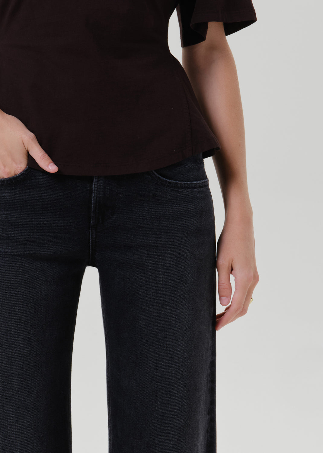 Close-up of a person wearing dark charcoal gray wide-leg jeans and a dark brown top, standing against a grey background.