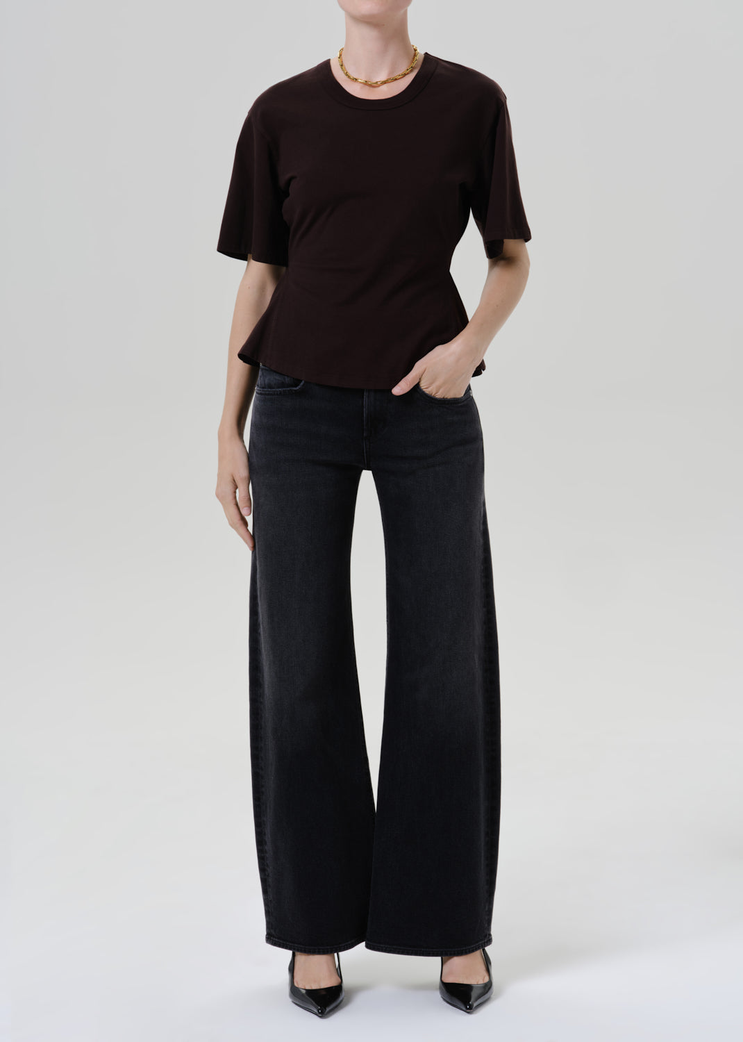 A model wearing a dark brown short-sleeve crewneck top and black wide-leg pants, standing against a grey background.
