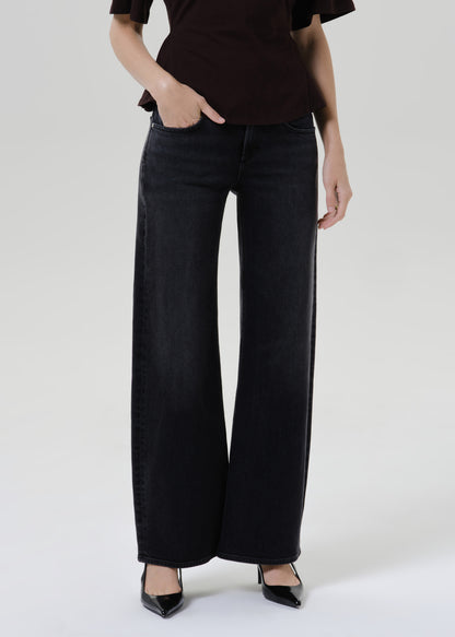 A model wearing black wide-leg denim jeans, paired with a dark brown top and black pointed-toe heels, standing against a grey background.