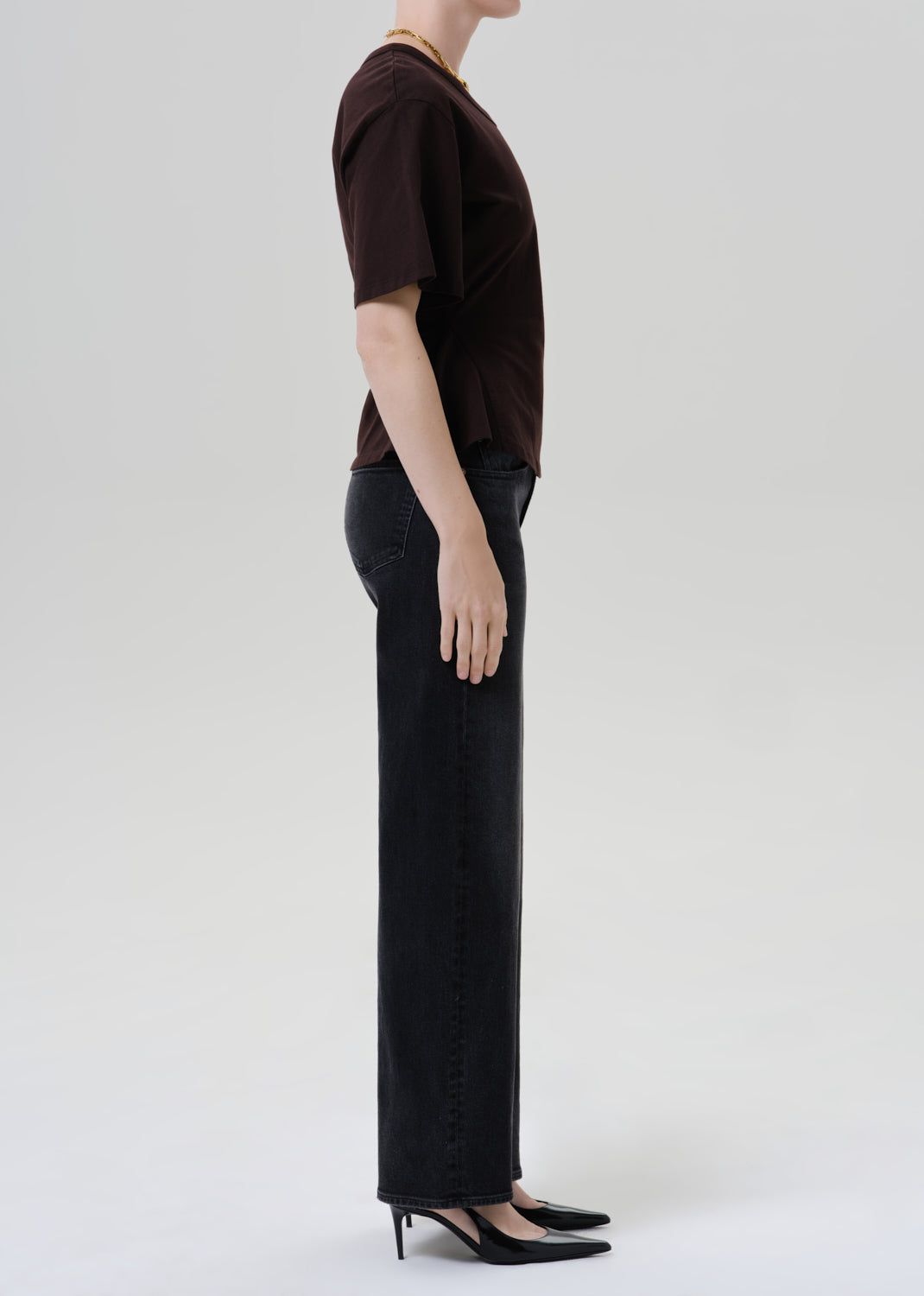 A side-profile studio shot of a model wearing a dark brown short-sleeve t-shirt, black wide-leg jeans, and black pointed-toe heels against a grey background.