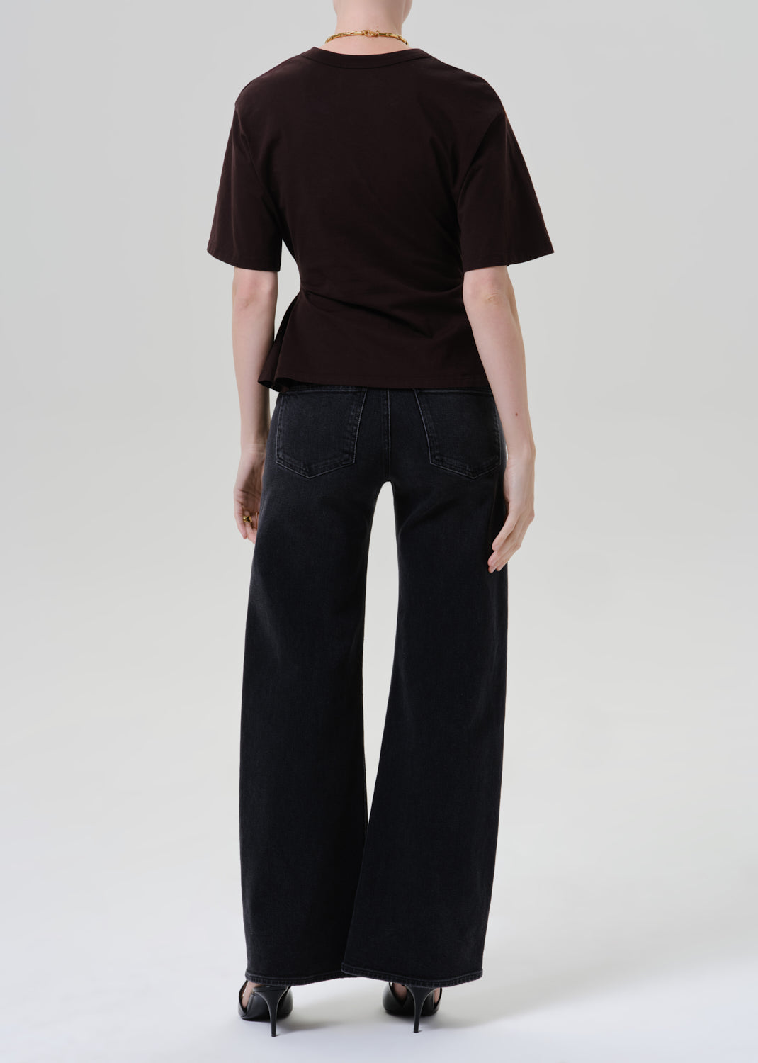 A model shown from the back wearing a dark brown short-sleeve top with a peplum waist and black wide-leg jeans, photographed against a grey background.