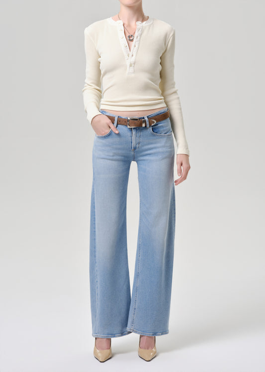 A model wearing a cream long-sleeve henley top with lace trim, light-wash wide-leg jeans, a brown belt, and beige heels, photographed against a grey background.