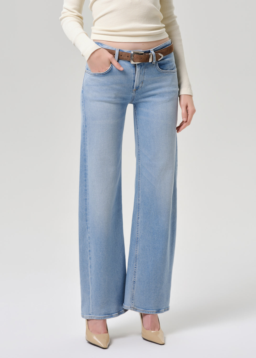 A model wearing light-wash wide-leg denim jeans with a brown belt, paired with a cream long-sleeve top and beige pointed-toe heels, photographed against a grey background.