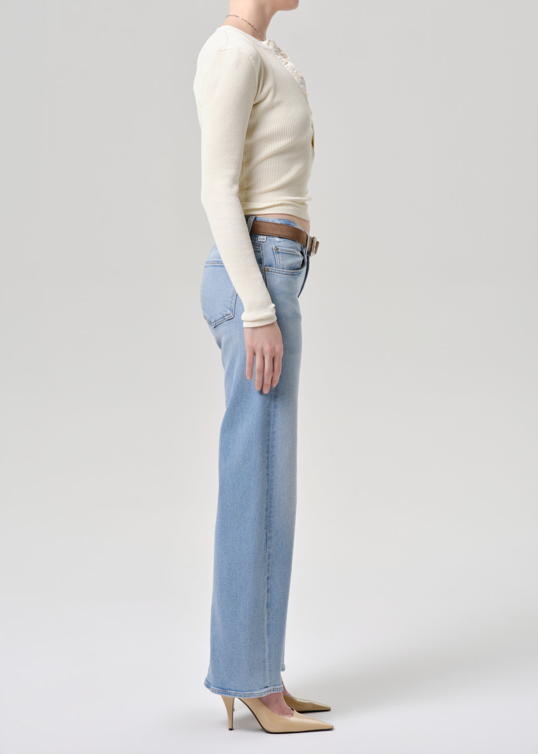 A model shown in profile wearing a cream ribbed long-sleeve top, light-wash wide-leg jeans, and beige pointed-toe heels against a grey background.