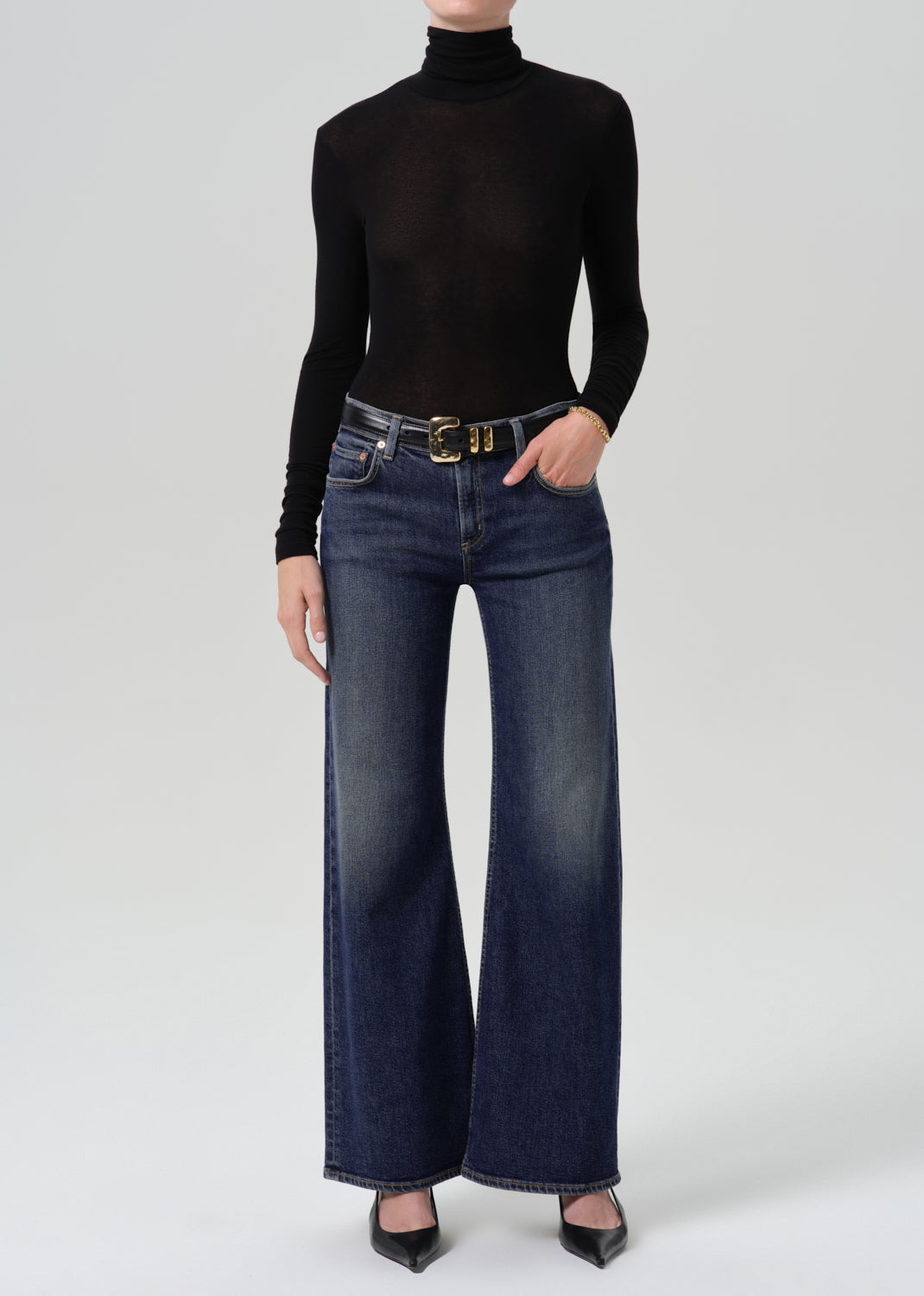 Person wearing dark blue wide leg jeans with subtle fading, a black long sleeve turtleneck top, and black pointed-toe heels, front full body view on a grey background.