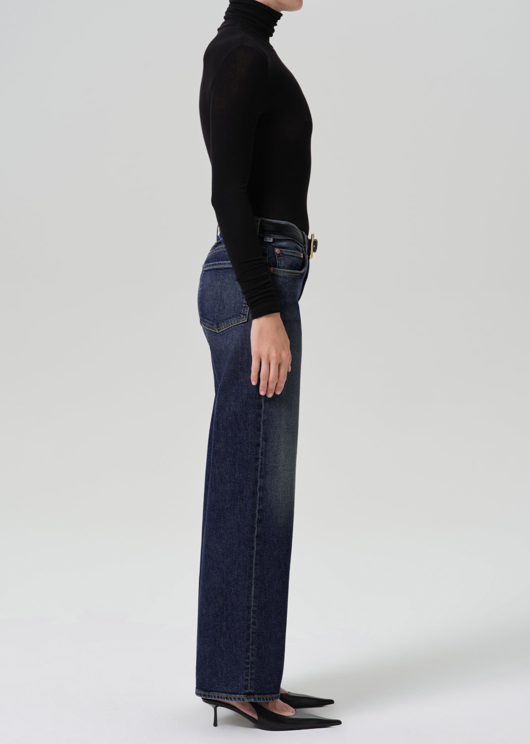 Person wearing dark blue wide leg jeans with a black long sleeve turtleneck top and black heels, side full body view on a grey background.
