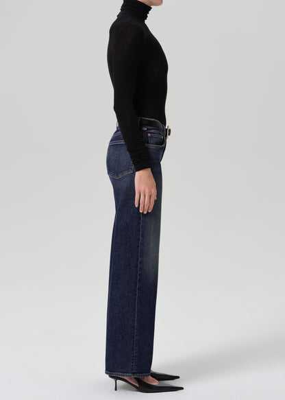 Person wearing dark blue wide leg jeans with a black long sleeve turtleneck top and black heels, side full body view on a grey background.
