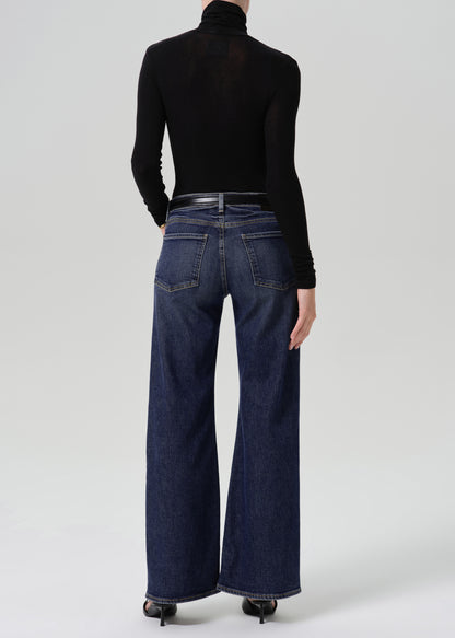 Person wearing dark blue wide leg jeans with a black long sleeve turtleneck top and black heels, back full body view on a grey background.
