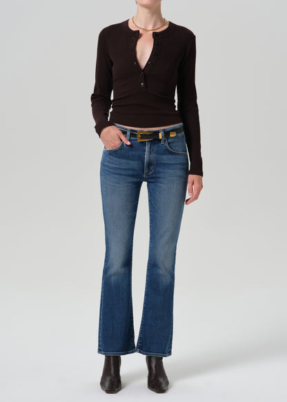 A model wearing a dark brown long-sleeve henley top, blue flare jeans with a gold-tone belt, and dark brown ankle boots, photographed against a grey background.