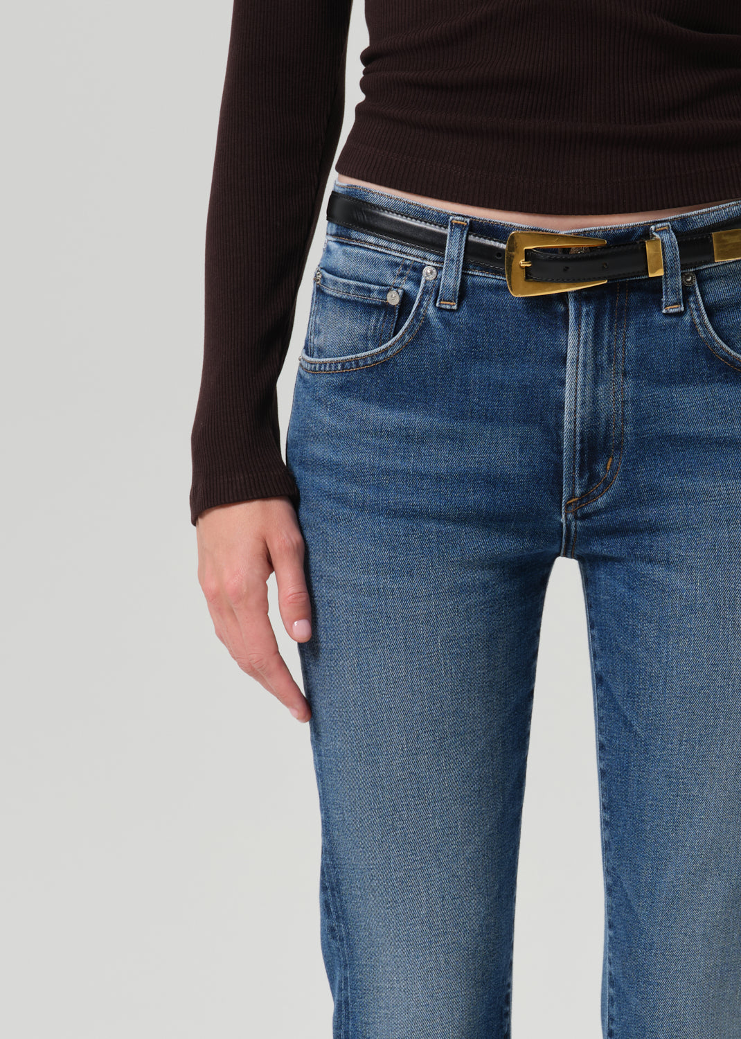 Close-up of a person wearing blue jeans with a black belt featuring a gold buckle, paired with a dark brown ribbed long-sleeve top, against a grey background.