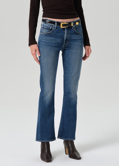 A model wearing a pair of medium-wash blue flare jeans with a gold-tone belt, paired with a dark brown long-sleeve top and dark brown heeled ankle boots, against a grey background.