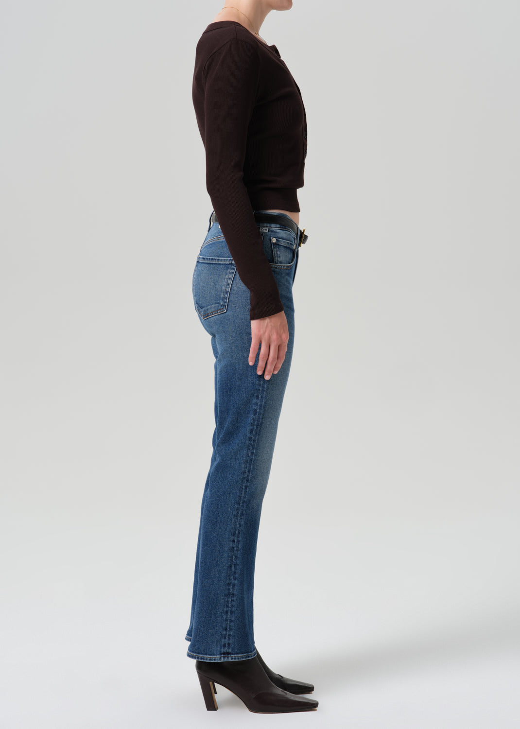 A side-profile studio shot of a model wearing blue straight-leg jeans, a dark brown long-sleeve crop top, and black heeled ankle boots against a grey background.