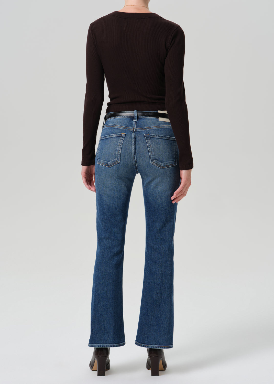 Rear view of a model wearing blue flare jeans, a dark brown long-sleeve top, and black heels against a grey background.