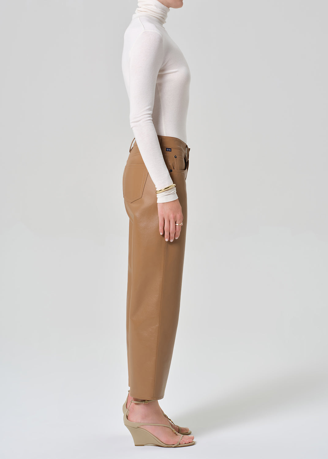 Person wearing a white turtleneck, beige sweater, and tan wide-leg leather pants on a grey background