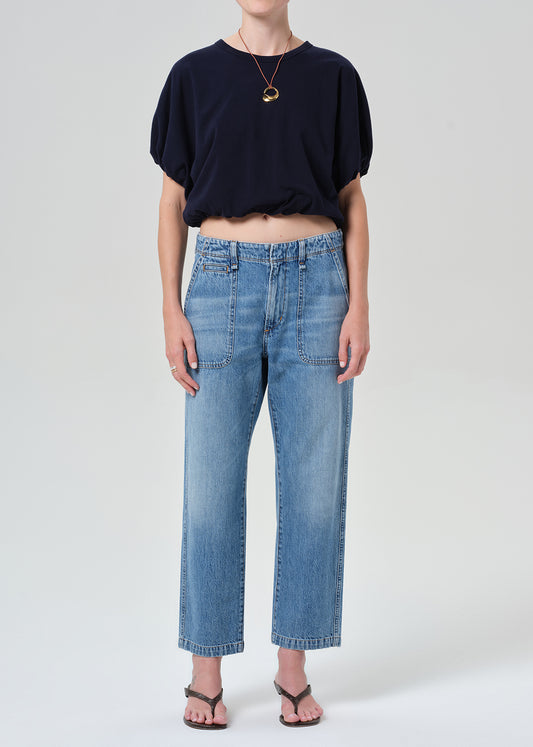 A model wearing a navy blue cropped short-sleeve top, light-wash blue cropped jeans, and brown thong sandals, standing against a grey background.