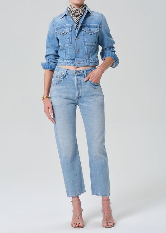 A model wearing a light-wash blue denim jacket and matching high-waisted light-wash blue denim jeans, styled with a black and white patterned scarf, gold bracelets, and nude strappy heels against a grey background.