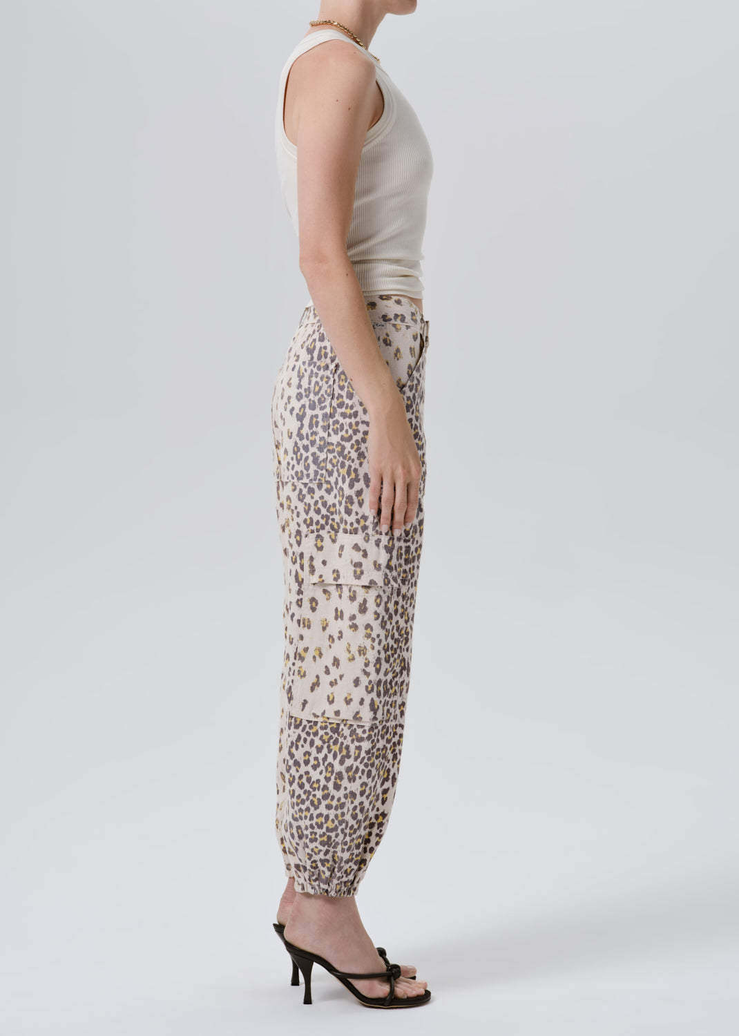 A side-profile view of a model wearing beige animal-print harem pants with a cream sleeveless top and black heeled sandals, set against a grey background.