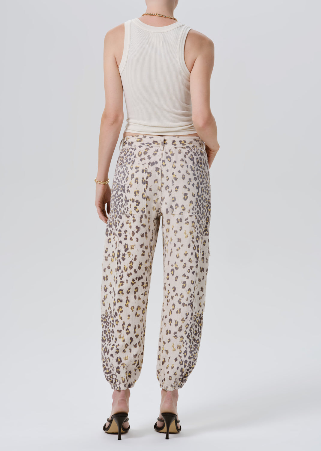 Rear view of a model wearing beige leopard-print pants with a cream tank top, standing against a grey background.