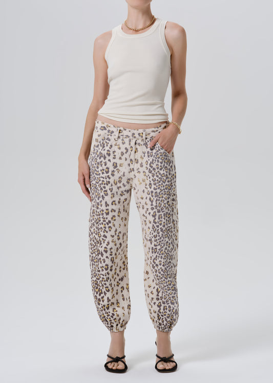 A model wearing beige leopard-print pants with a cream ribbed tank top and black sandals, standing against a grey background.