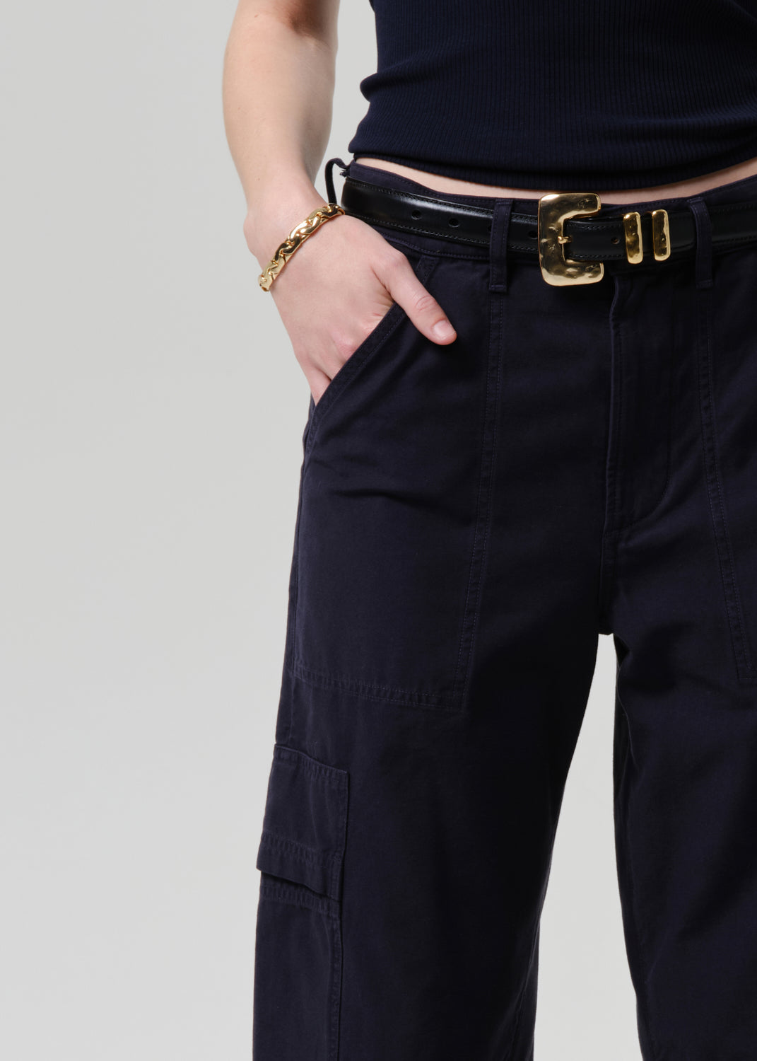 Person wearing black cargo pants with a gold belt buckle on a grey background