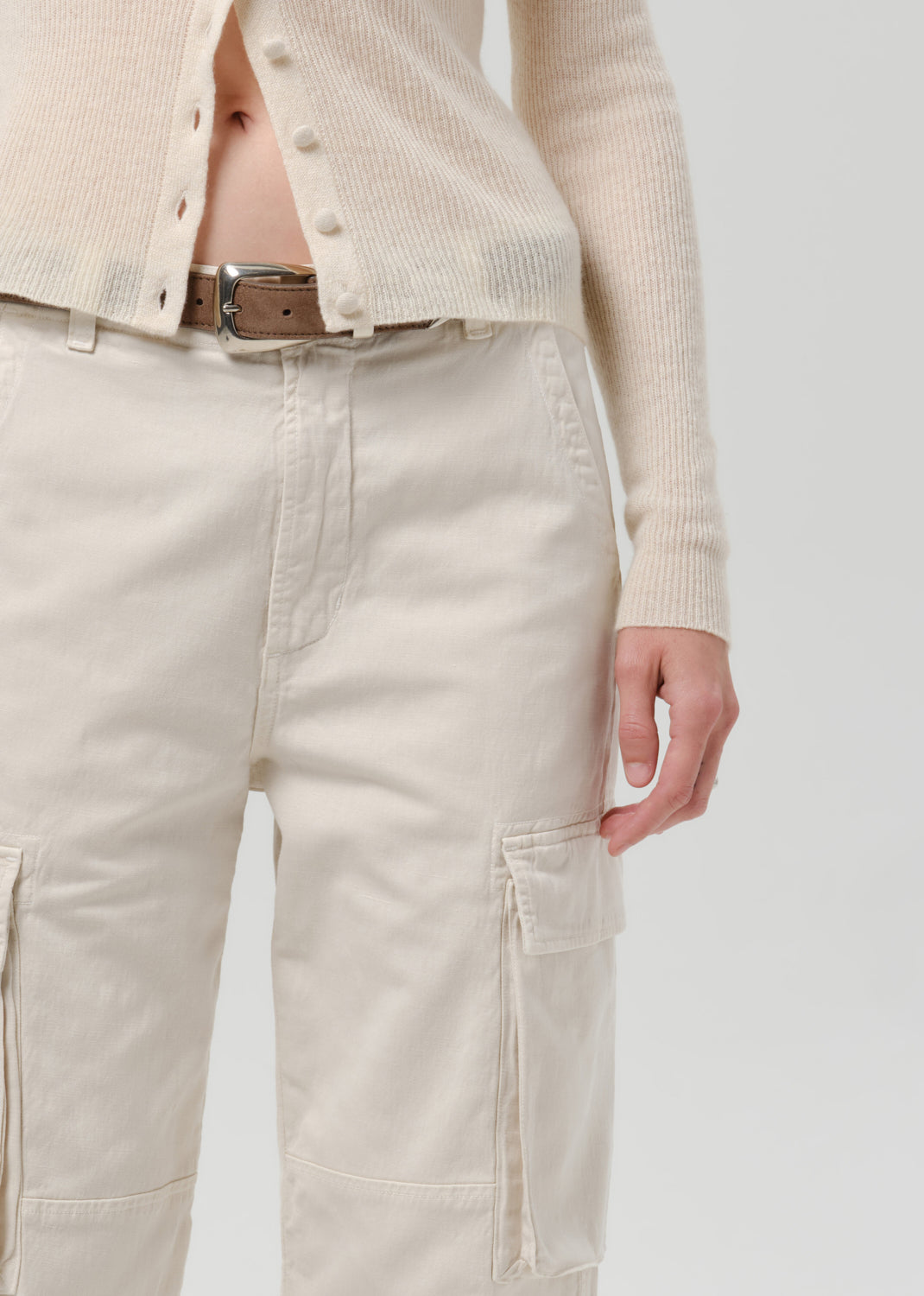 Close-up of a person wearing off-white cargo pants with a brown belt and a matching off-white ribbed cardigan, against a grey background.