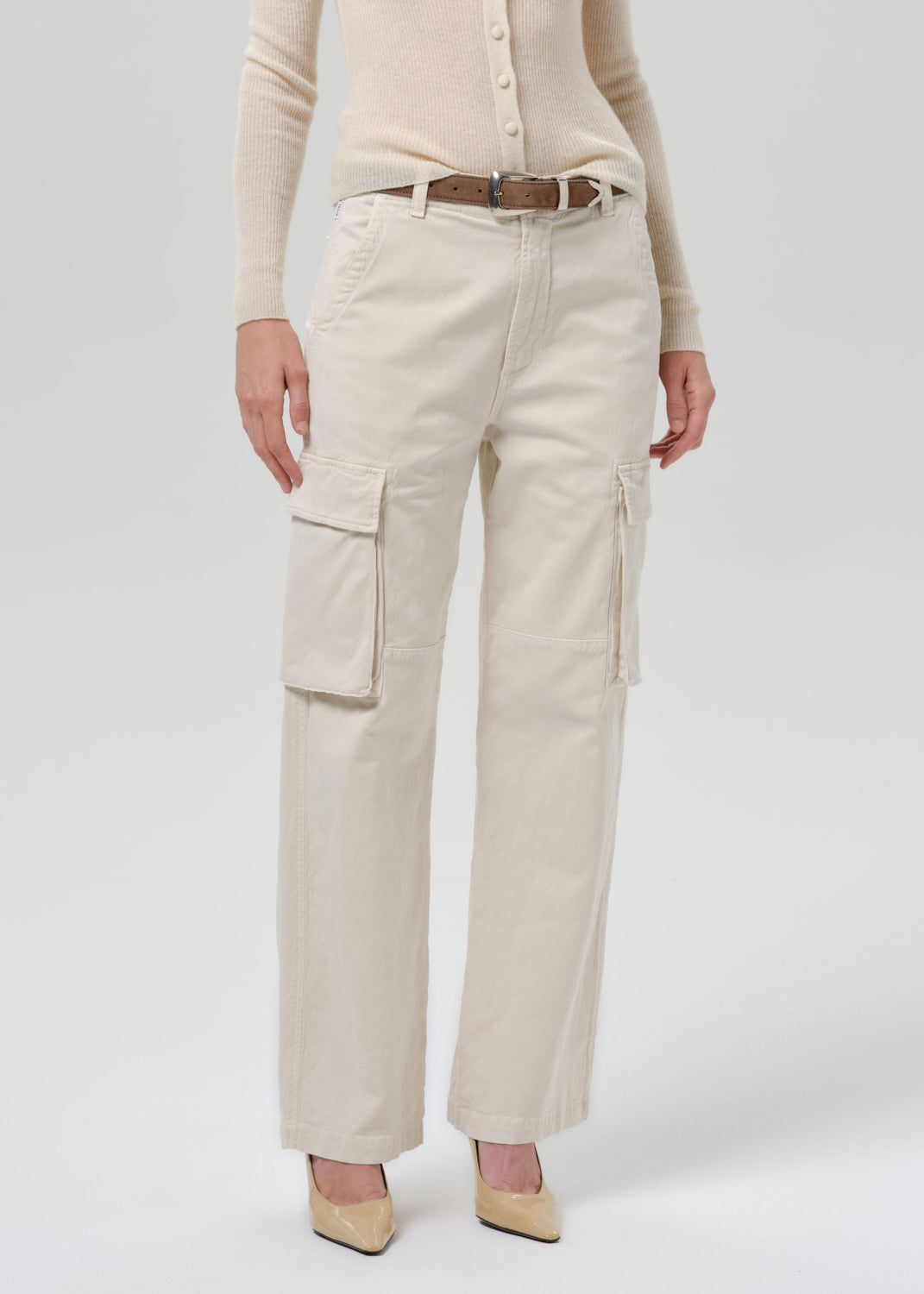 A model wearing a pair of off-white cargo pants with a brown belt, paired with a beige cardigan and beige heels, shown from the waist down on a grey  background.
