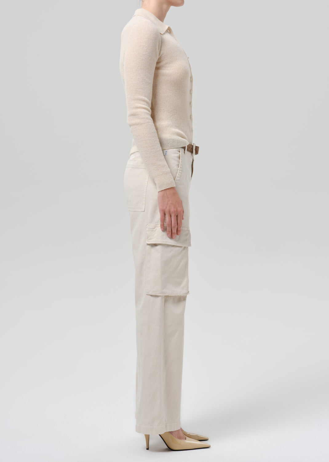 A model shown in profile wearing a cream-colored long-sleeve knit top and matching high-waisted cargo pants with a brown belt, paired with beige pointed-toe heels, against a grey background.