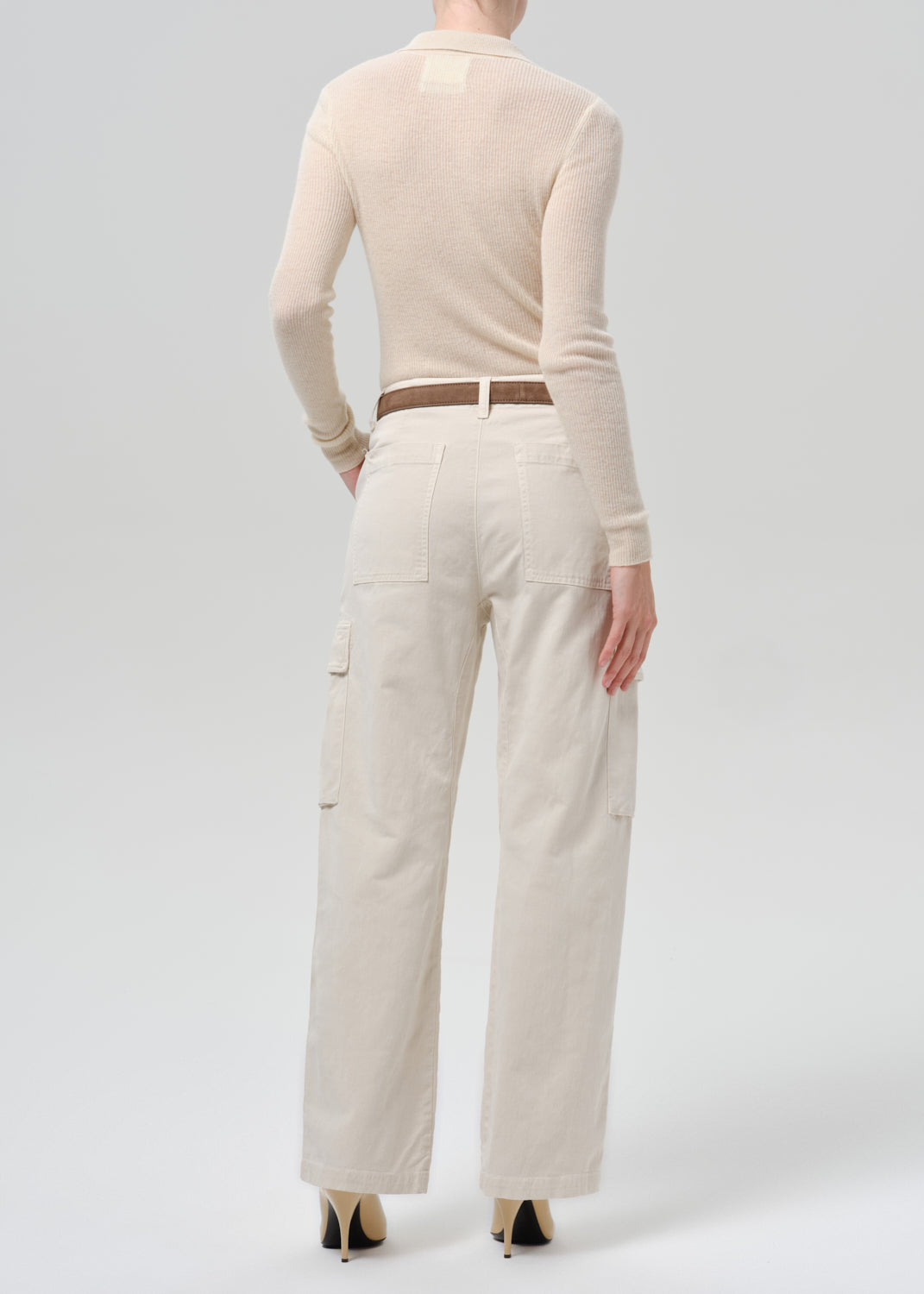 A model shown from the back wearing a cream-colored long-sleeve knit top and matching wide-leg cargo pants, styled with a brown belt and beige high heels against a grey background.