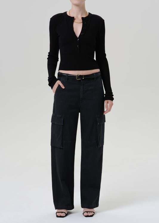 A model wearing a black long-sleeve cropped henley top and black wide-leg cargo pants with a black belt, standing against a grey background.