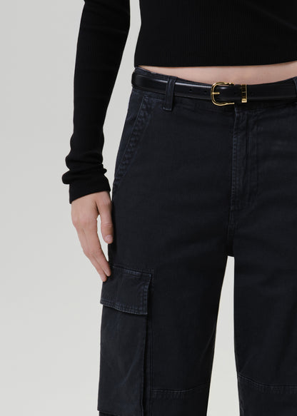 Close-up of a person wearing dark cargo pants with a black belt and gold buckle, paired with a black long-sleeve top, against a grey background.