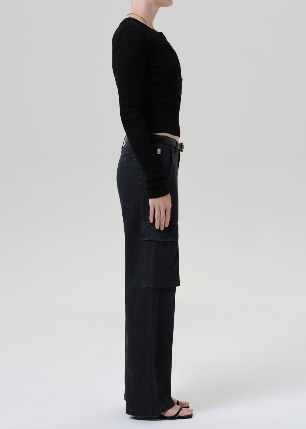 A side-profile studio shot of a model wearing a black long-sleeve crop top and black wide-leg cargo pants against a grey background.