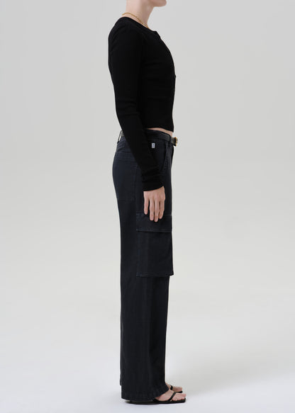A side-profile studio shot of a model wearing a black long-sleeve crop top and black wide-leg cargo pants against a grey background.
