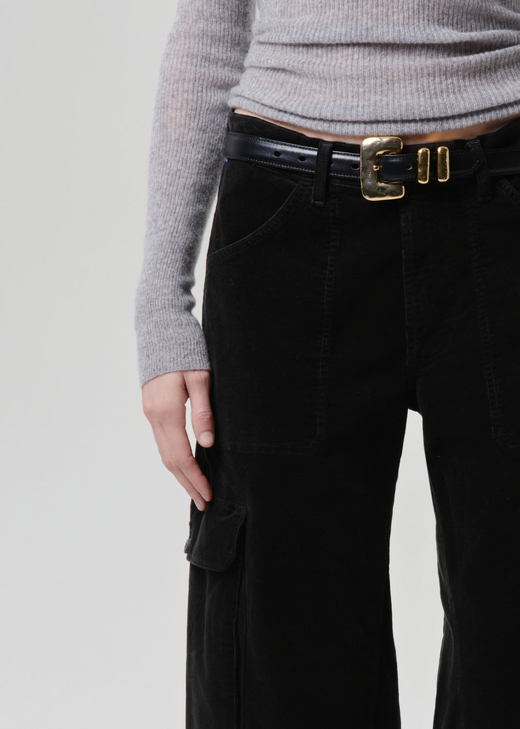 Person wearing dark green pants with a gold belt buckle on a grey background