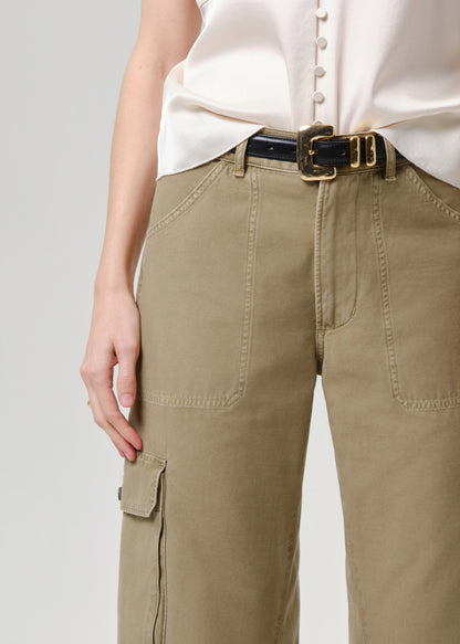 A close-up of a person wearing khaki cargo pants with a black leather belt featuring a gold buckle, paired with a cream satin blouse, against a grey background.