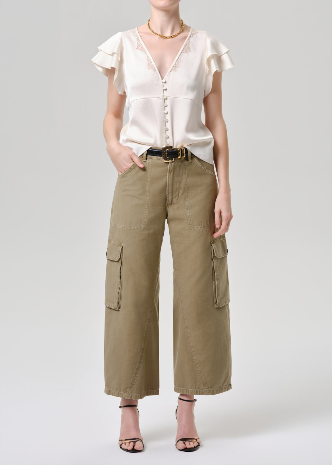 A model wearing a cream ruffled short-sleeve blouse with a button front and wide-leg olive cargo pants with side flap pockets, styled with a black belt and black strappy heels against a grey background.