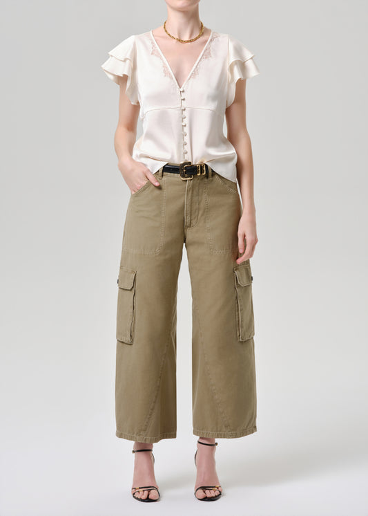 A model wearing a cream ruffled short-sleeve blouse with a button front and wide-leg olive cargo pants with side flap pockets, styled with a black belt and black strappy heels against a grey background.