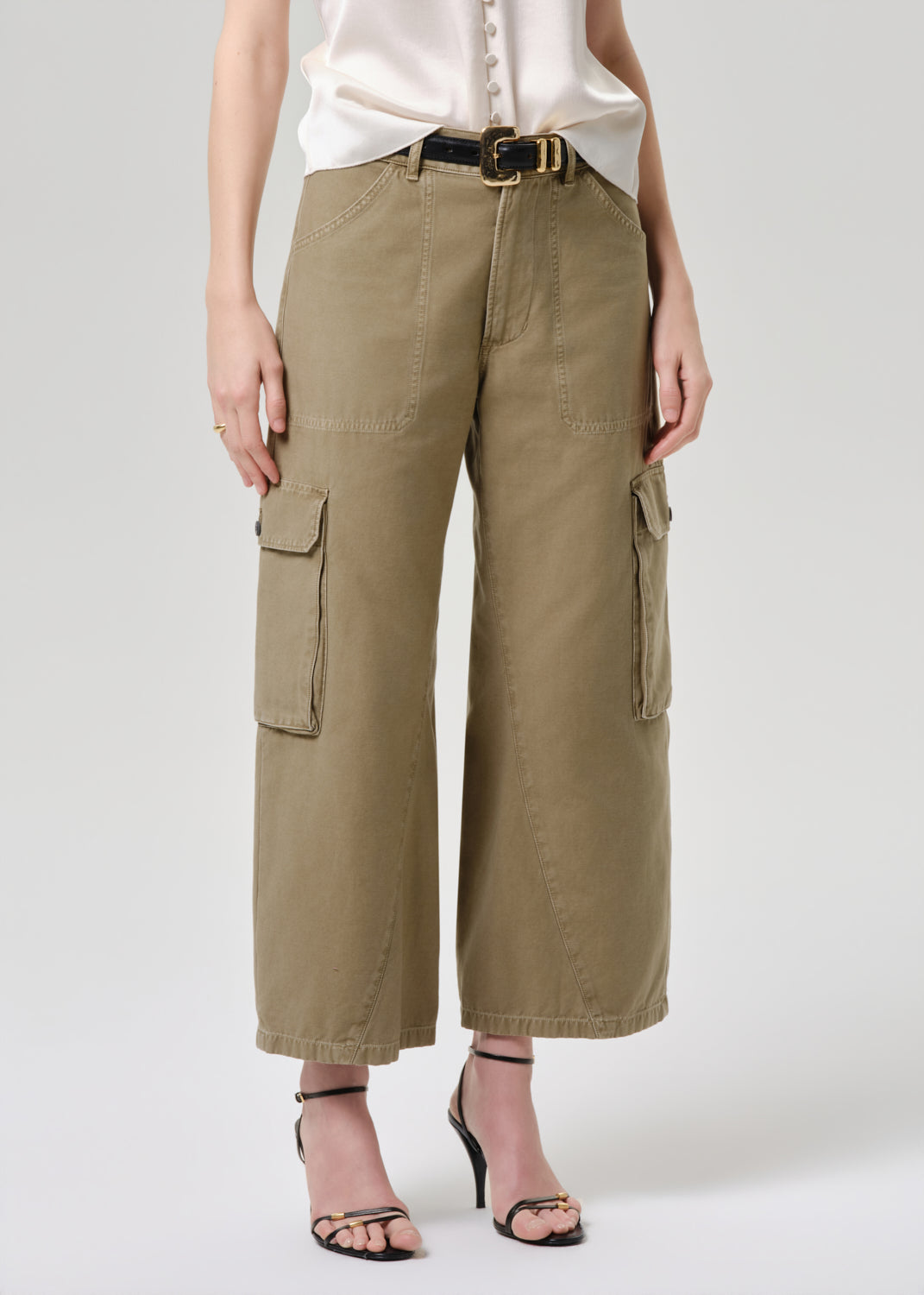 A model wearing olive green cropped cargo pants with a black belt, paired with a cream blouse and black strappy heels, against a grey background.