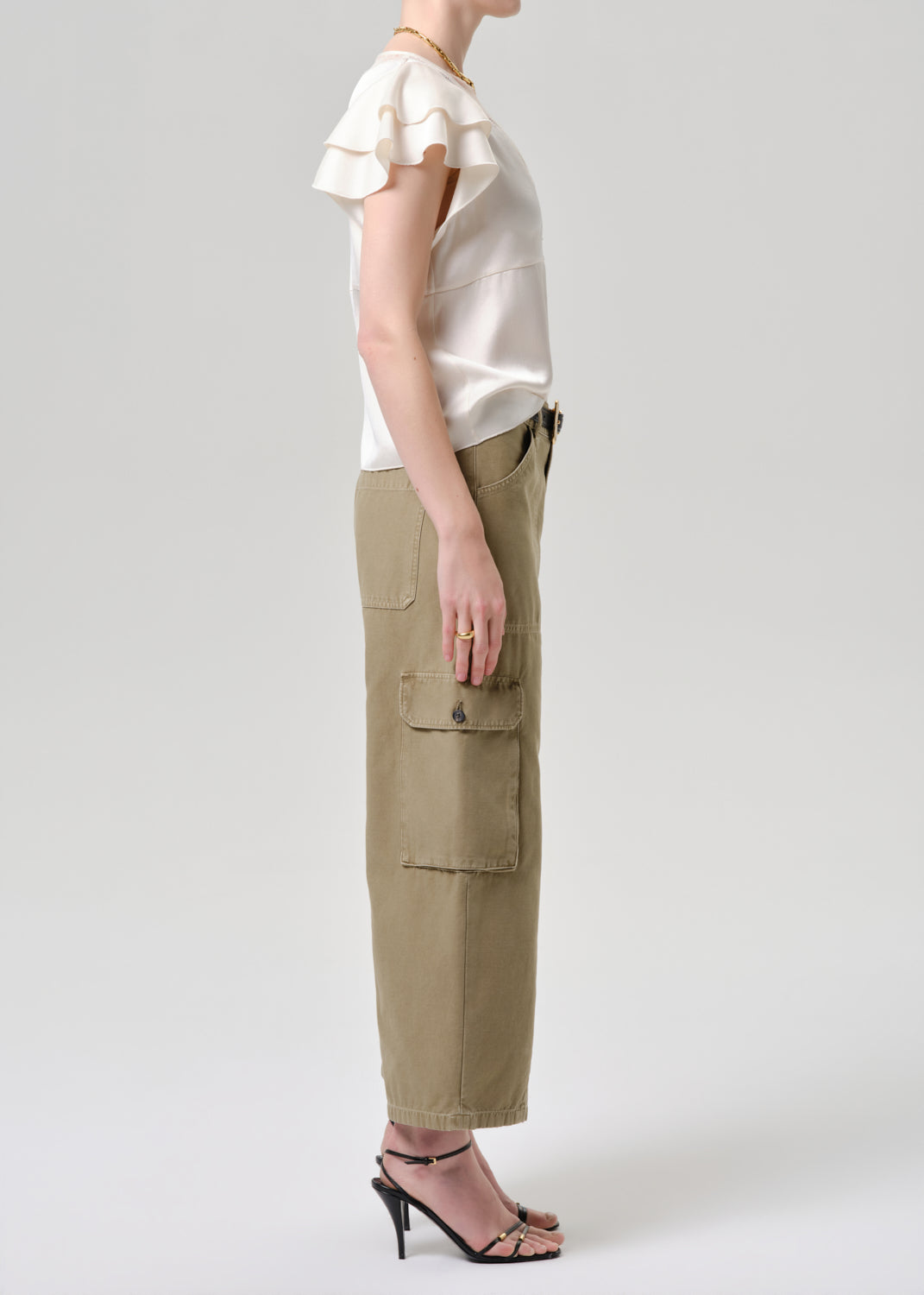 A model shown from the side wearing a cream ruffled short-sleeve top and olive green cargo pants with black high-heeled sandals, against a grey background.