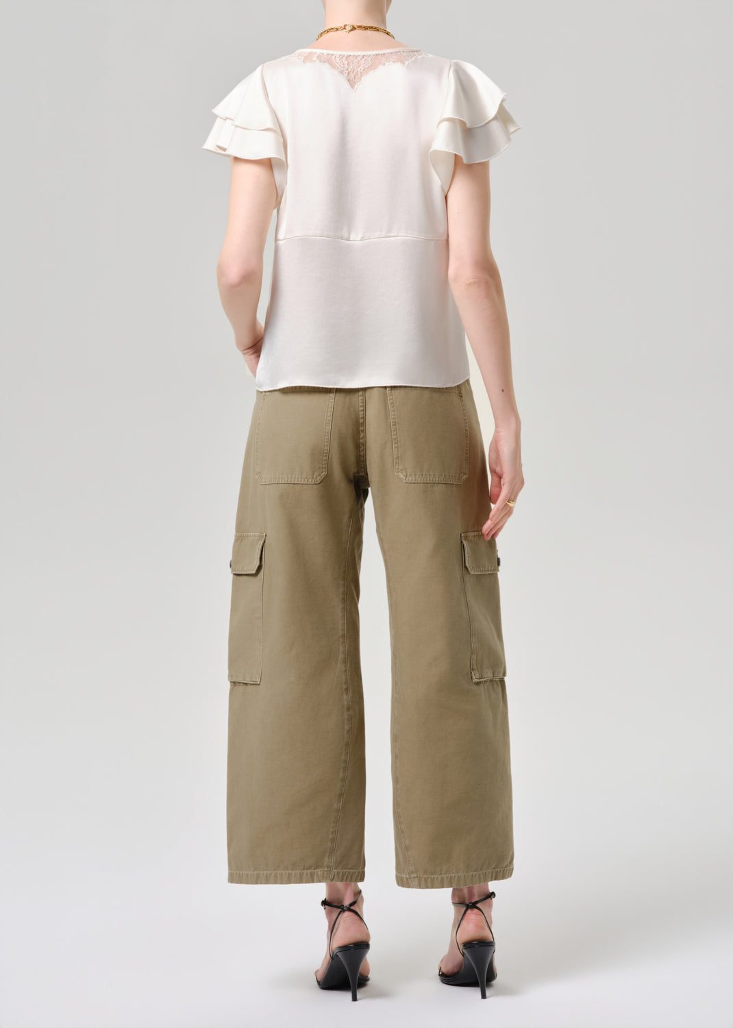A model shown from the back wearing a cream short-sleeve blouse with ruffle details and olive green wide-leg cargo pants, paired with black high-heeled sandals, against a grey background.