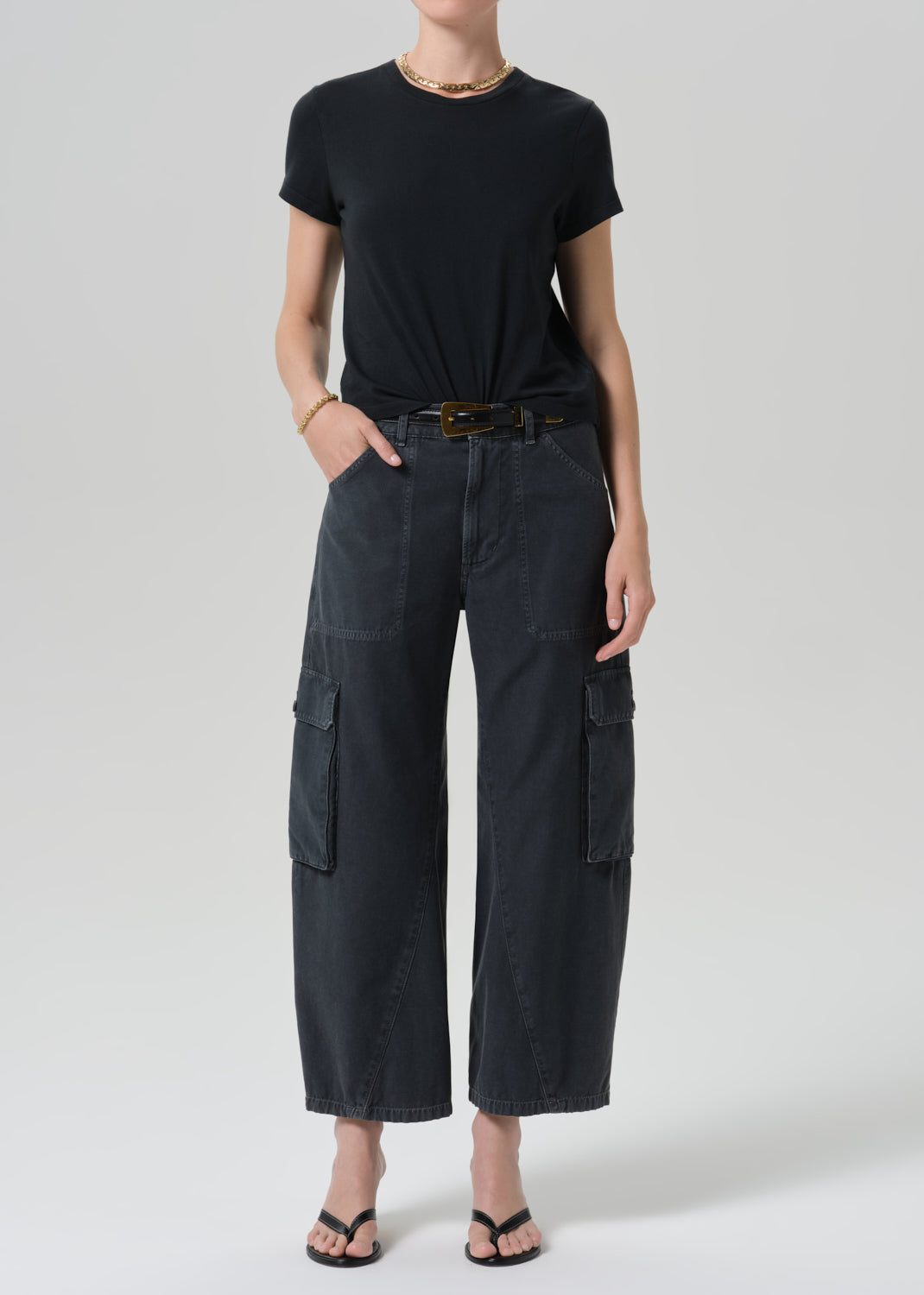 A model wearing a black short-sleeve t-shirt and dark charcoal gray wide-leg cargo pants with a black belt, styled with black sandals and a gold necklace against a grey background.
