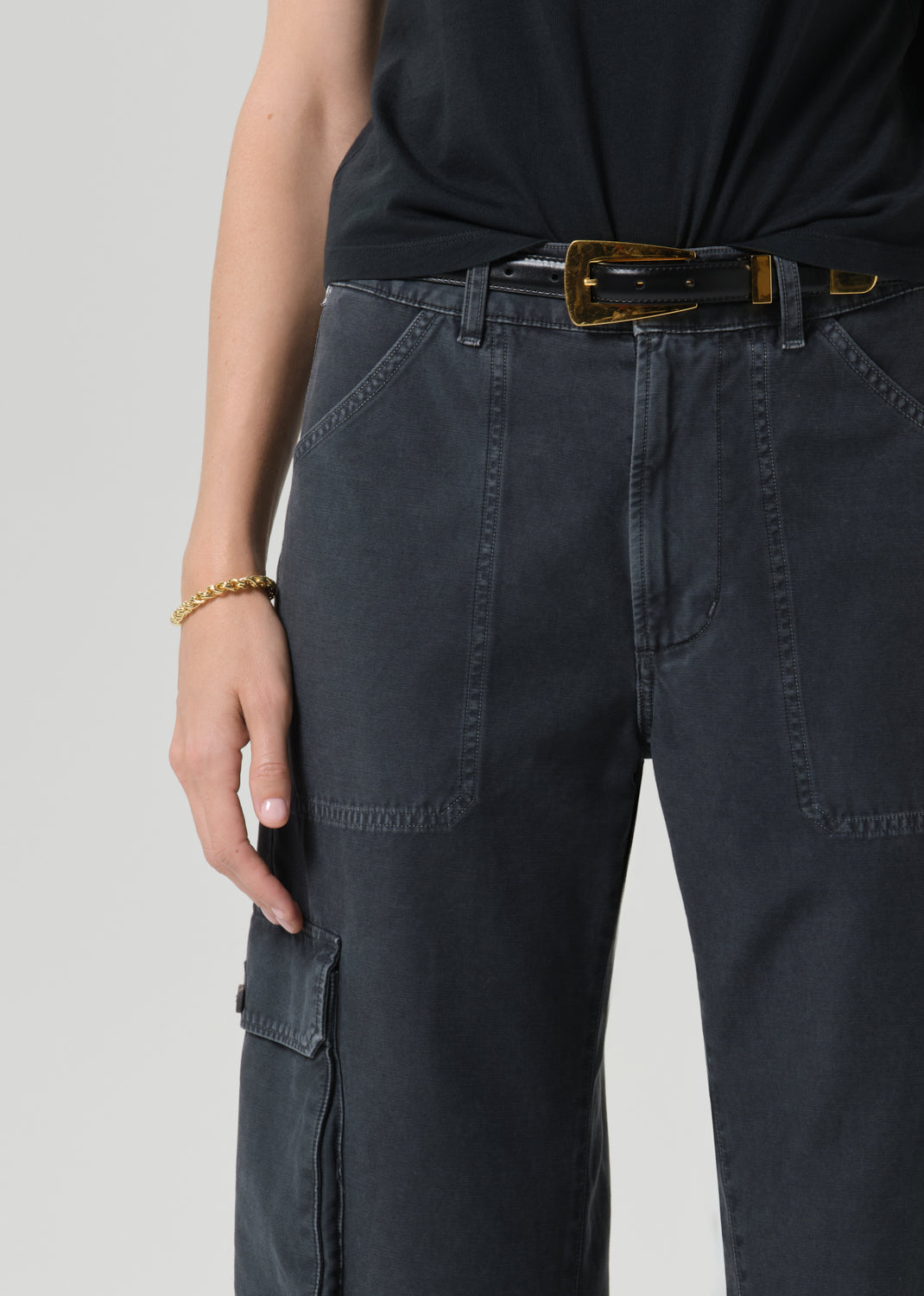 Close-up of a person wearing dark charcoal cargo pants with a black top and a gold-brown belt, shown against a grey background.