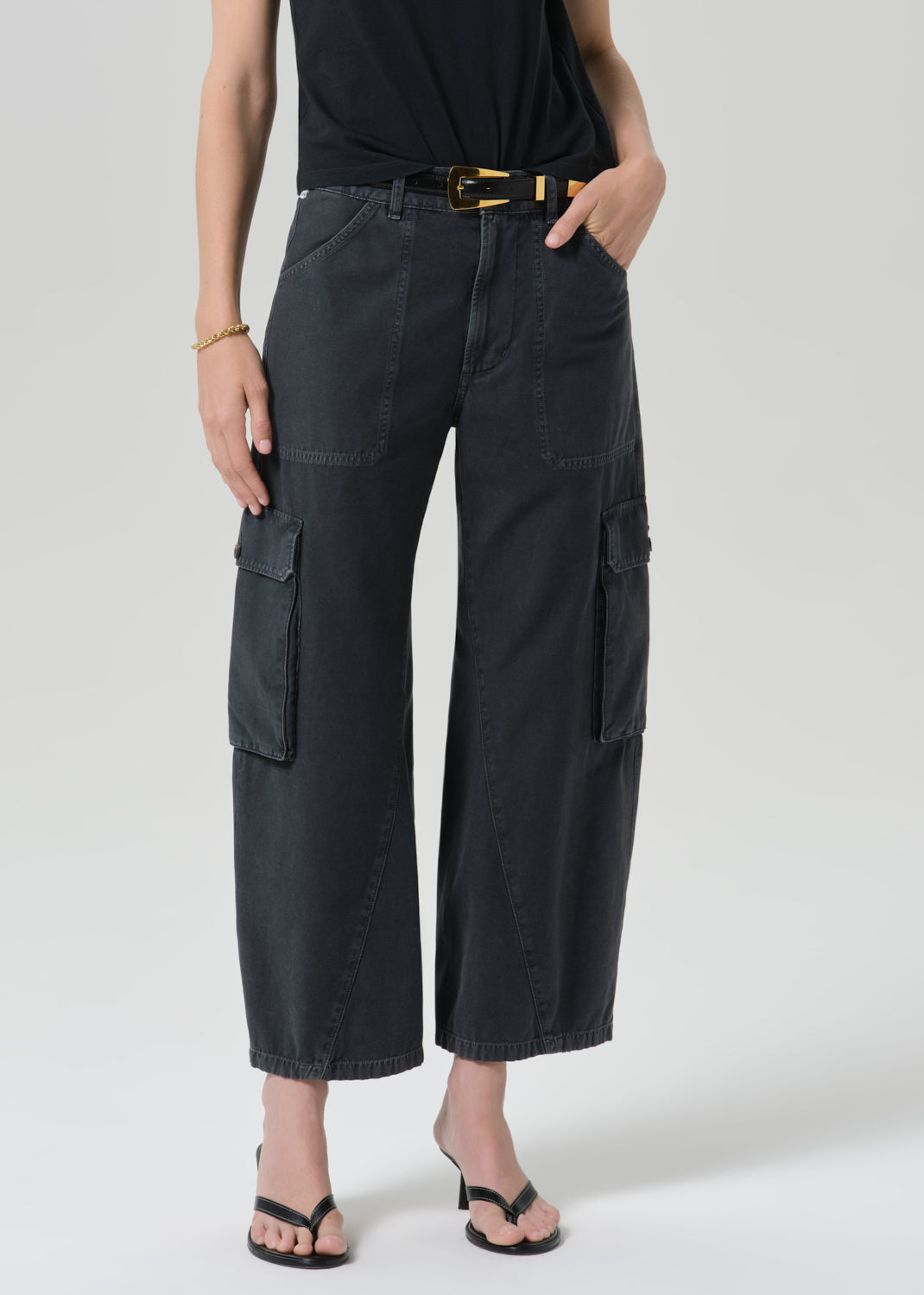 A model wearing black wide-leg cropped cargo pants with a black top, a gold-buckled belt, and black sandals, shown from the waist down against a grey background.