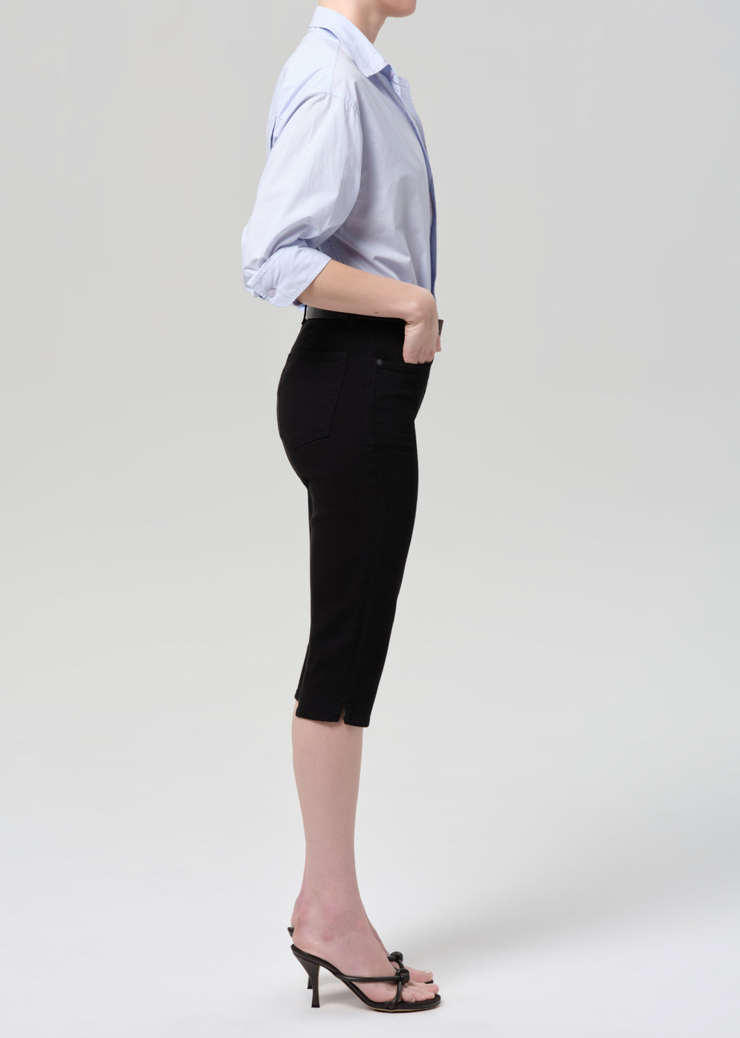 Person wearing a light blue shirt with rolled-up sleeves and black capris on a grey background