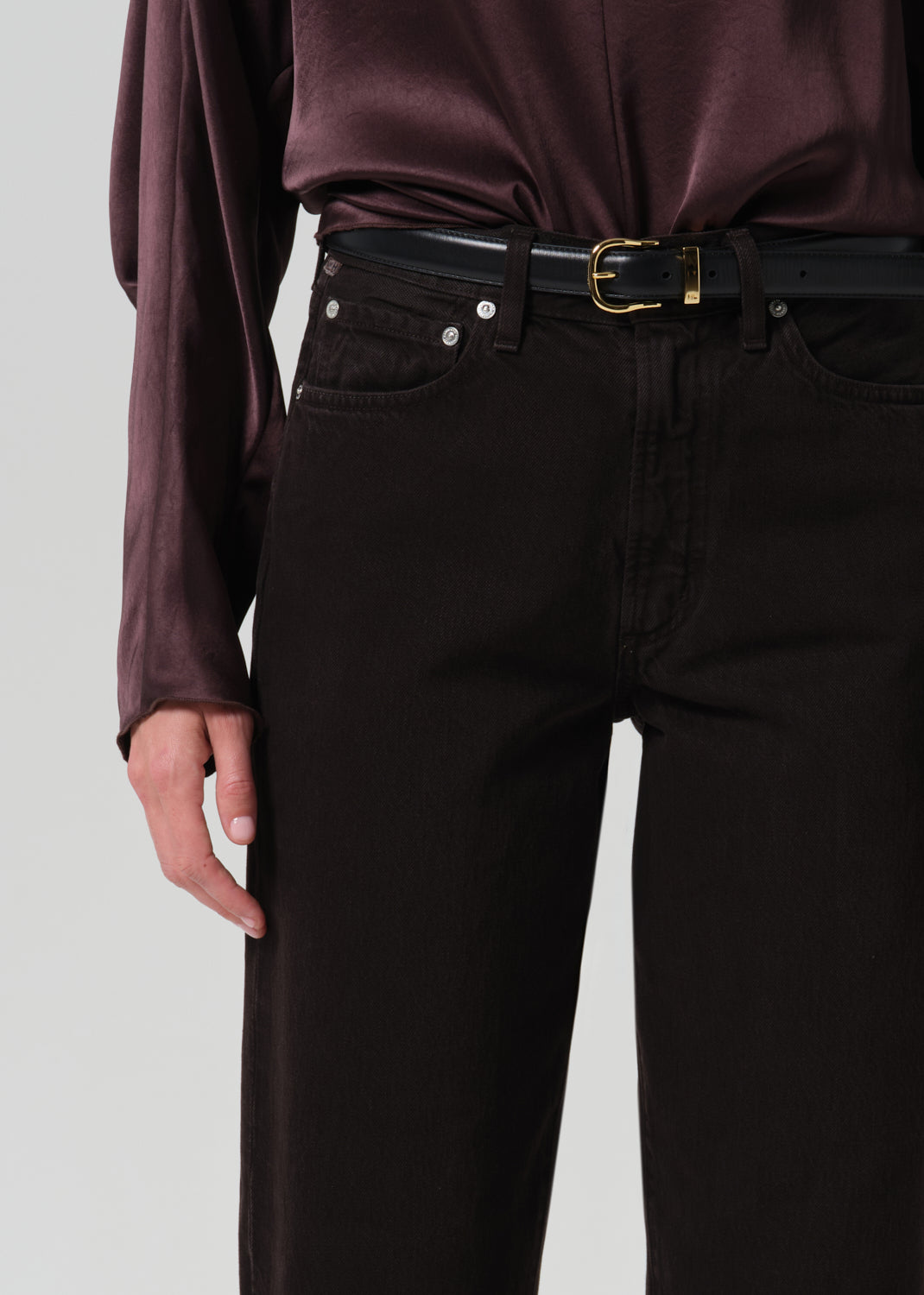 Close-up of a person wearing dark brown or near-black straight-leg jeans with a black belt and gold buckle, paired with a dark plum satin blouse, against a grey background.
