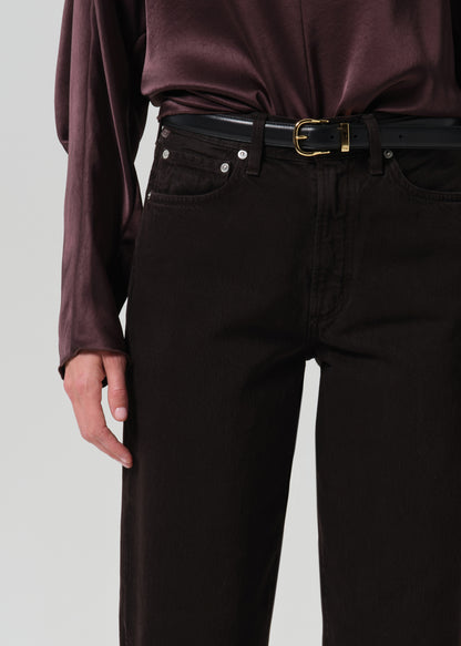 Close-up of a person wearing dark brown or near-black straight-leg jeans with a black belt and gold buckle, paired with a dark plum satin blouse, against a grey background.