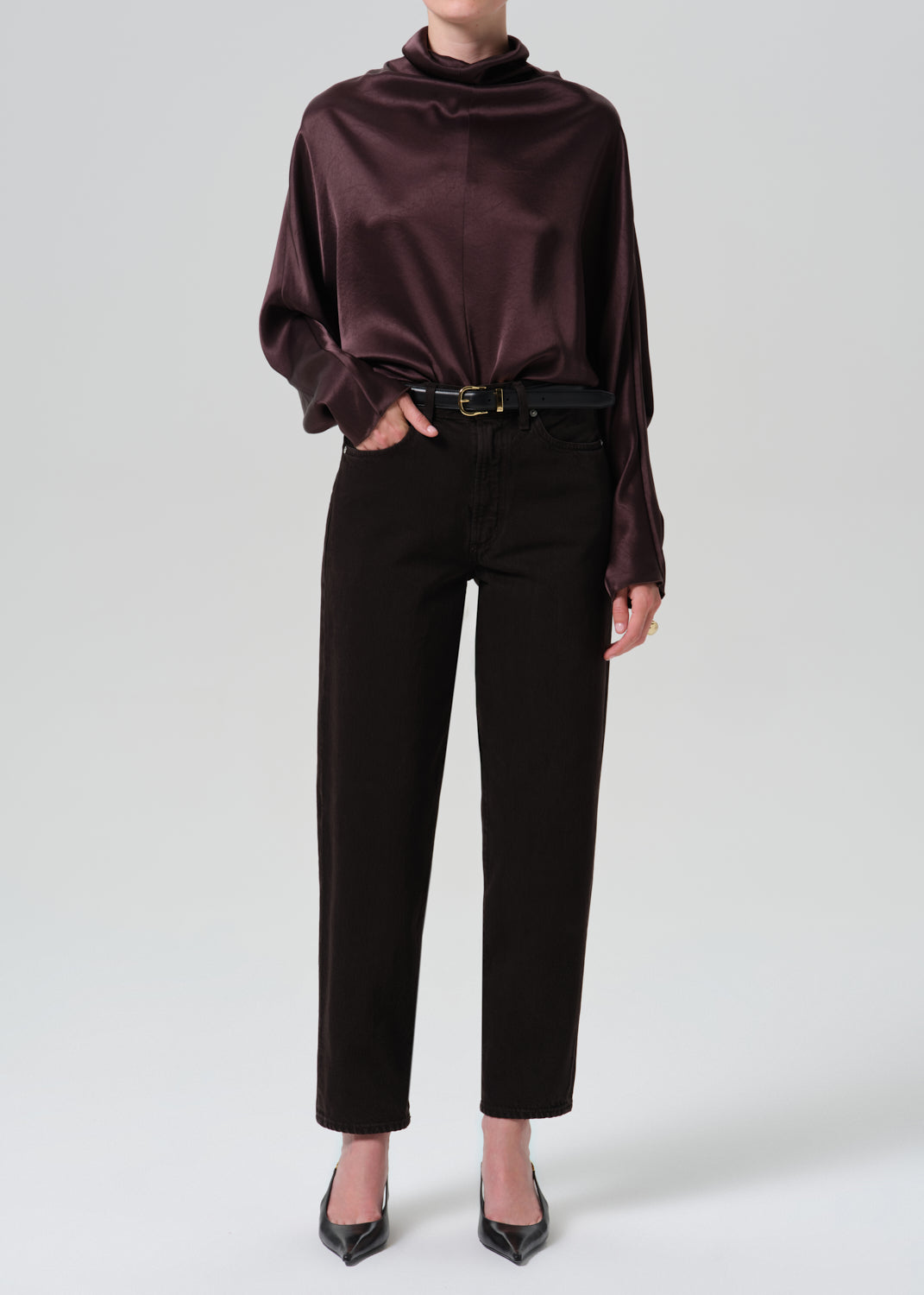 A model wearing a dark plum satin long-sleeve top, black trousers, and black pointed-toe heels, photographed against a grey background.