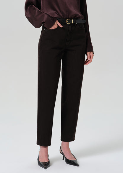 A model wearing dark brown slim-fit cropped pants with a black belt, paired with a dark brown satin blouse and black pointed-toe heels, shown from the waist down on a grey background.