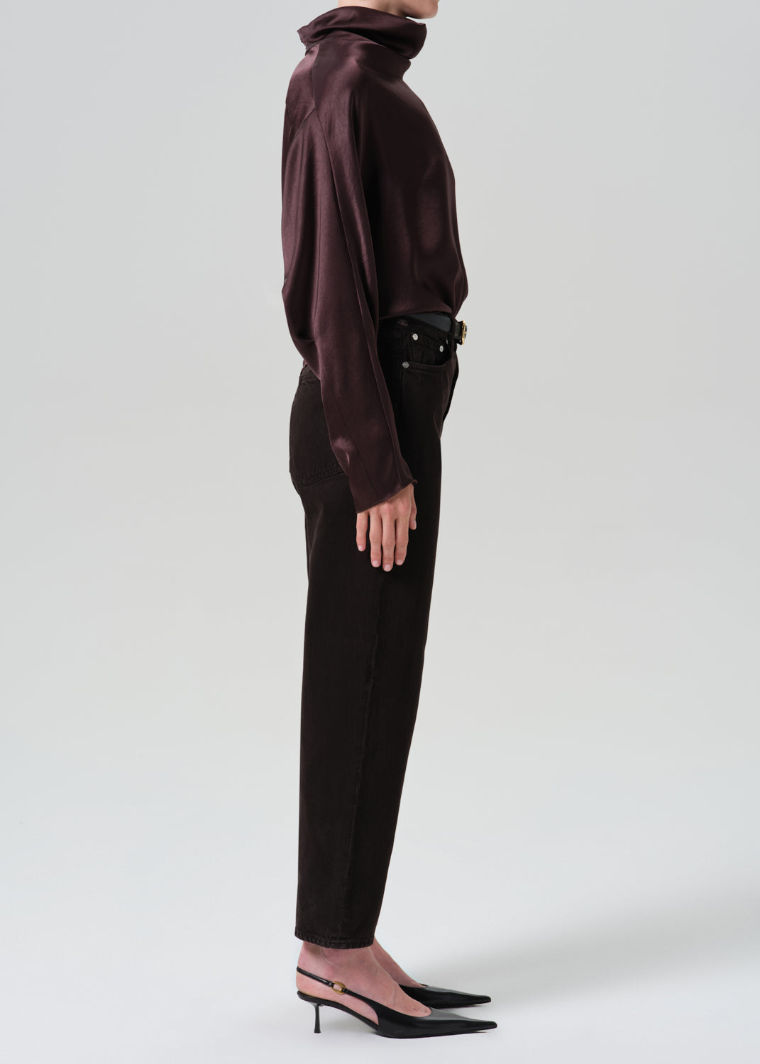 A model shown in profile wearing a dark plum satin long-sleeve turtleneck top, dark brown trousers, a black belt, and black pointed-toe slingback heels against a grey background.